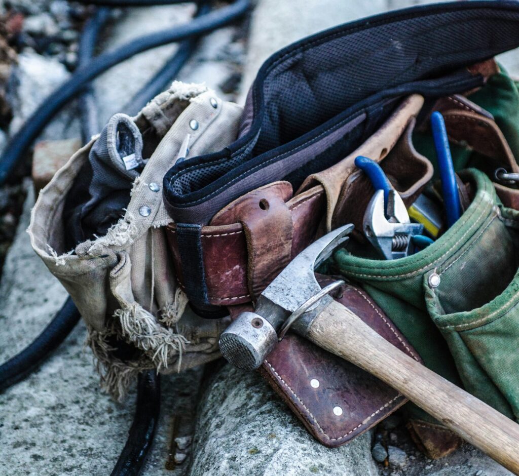 Tool belt filled with various hand tools, including a hammer, pliers, and screwdrivers, resting on a concrete surface with black hoses in the background.