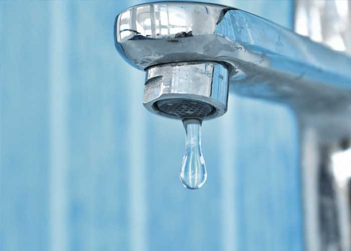 Close-up image of a chrome faucet with a single water droplet hanging from the spout, set against a light blue tiled background.